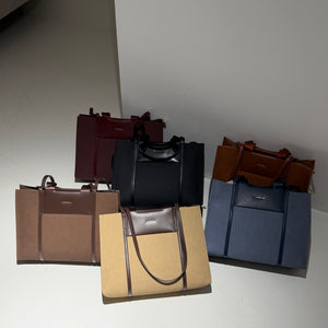 Four structured tote bags in different colors are arranged together on a light gray floor against a white wall. The bags include burgundy, black, tan with dark brown trim, and slate blue with dark blue trim.