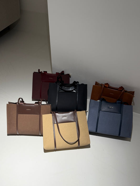 Four structured tote bags in different colors are arranged together on a light gray floor against a white wall. The bags include burgundy, black, tan with dark brown trim, and slate blue with dark blue trim.