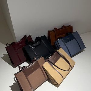 Six structured leather tote bags in assorted colors including burgundy, black, brown, tan, and navy, arranged on a white surface against a light gray wall.