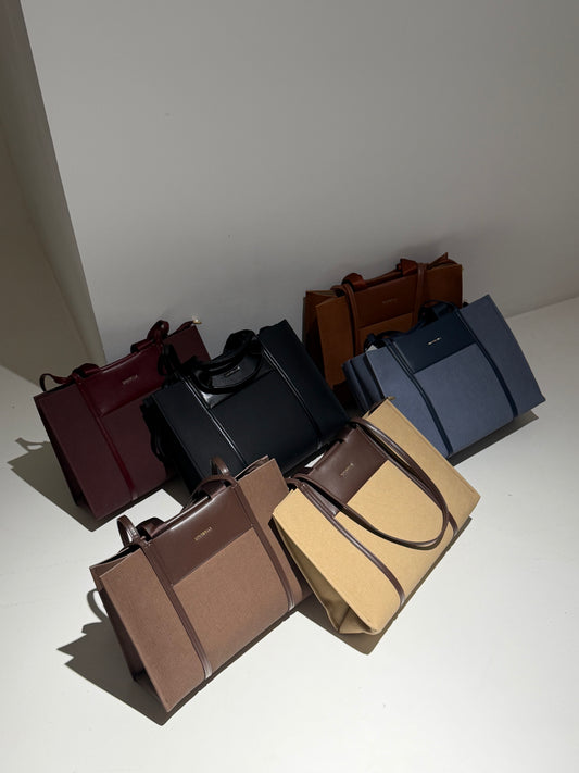 Six structured leather tote bags in assorted colors including burgundy, black, brown, tan, and navy, arranged on a white surface against a light gray wall.
