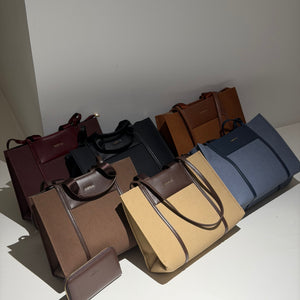 A collection of tote bags in multiple colors including burgundy, black, brown, tan, and blue, arranged on a white surface against a plain white wall.