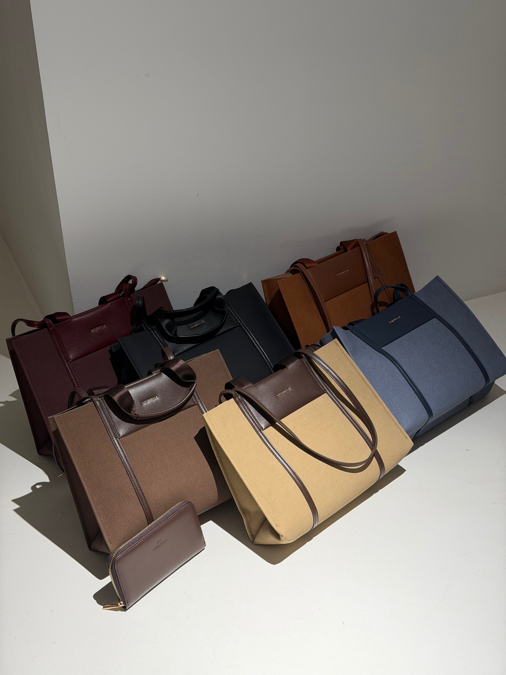 A collection of tote bags in multiple colors including burgundy, black, brown, tan, and blue, arranged on a white surface against a plain white wall.