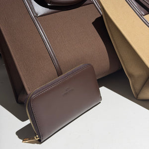 A brown leather zip-around wallet with gold-tone hardware and a subtle logo embossing, displayed in front of a matching brown fabric tote bag on a white surface with strong sunlight and shadows.