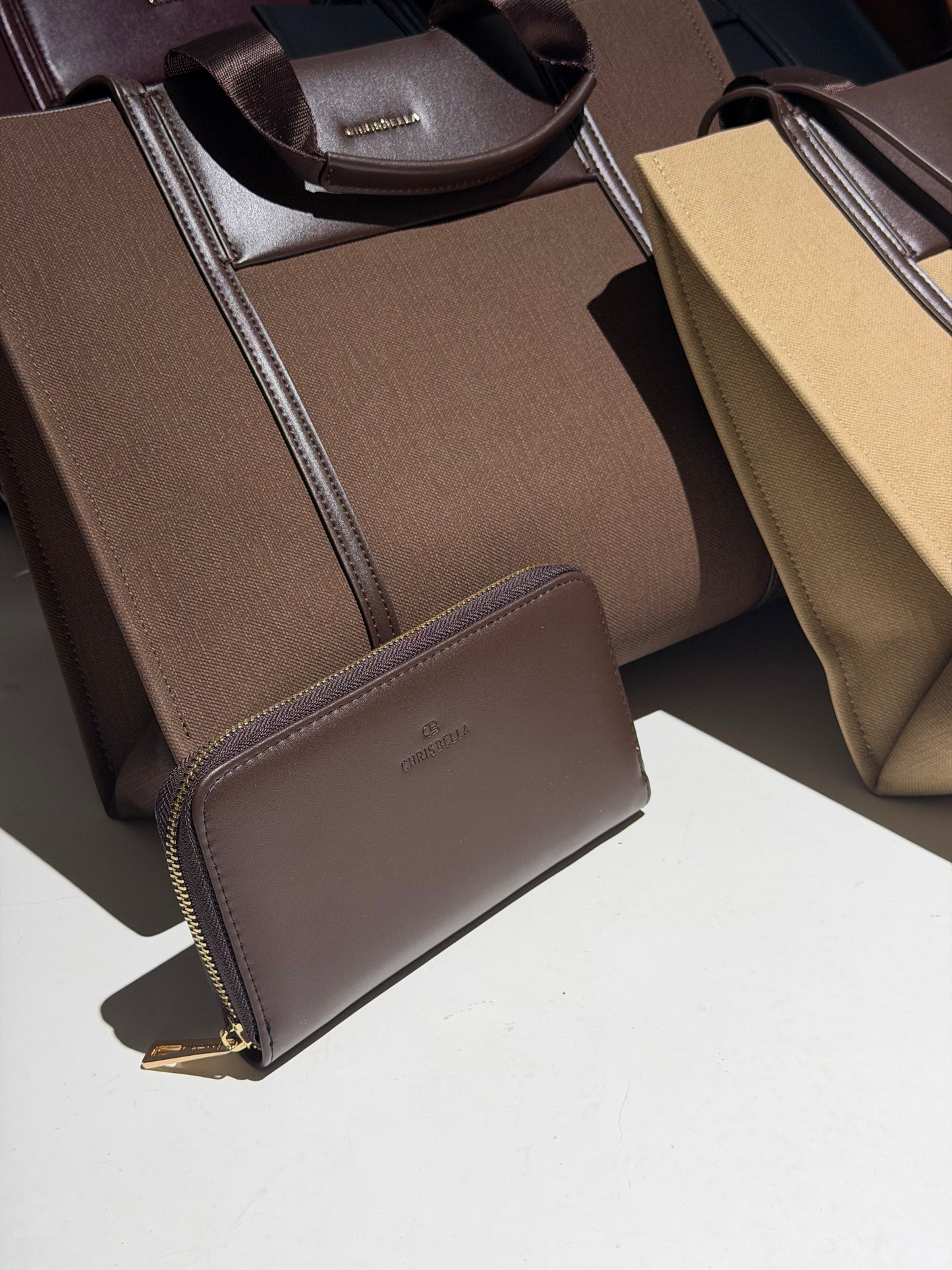 A brown leather zip-around wallet with gold-tone hardware and a subtle logo embossing, displayed in front of a matching brown fabric tote bag on a white surface with strong sunlight and shadows.